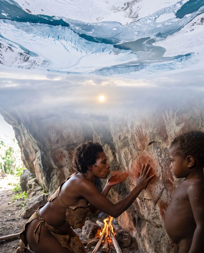 A woman blowing red ochre around her hand on a cave wall to make a hand stencil, a child watching.