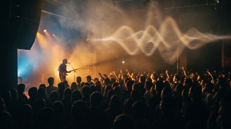 A musician on a small stage in a packed venue, a wave of light moving over the crowd.